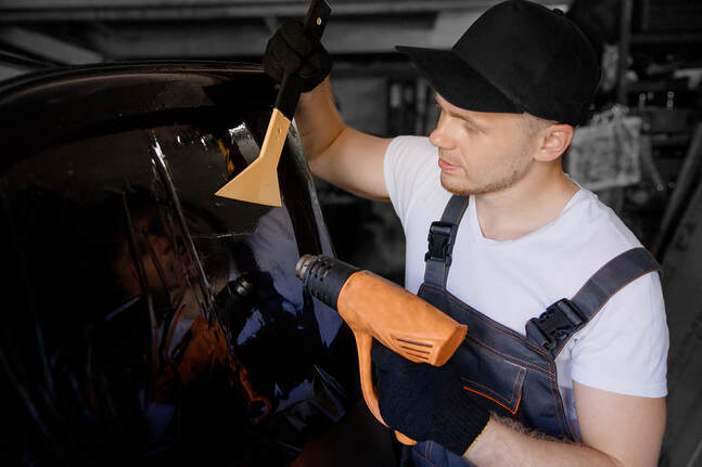 Picture of a guy holding an orange heat gun and scraper, while looking at the car