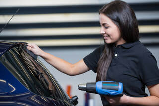 Picture of a blue car with a woman holding a blue heat gun while looking at the car